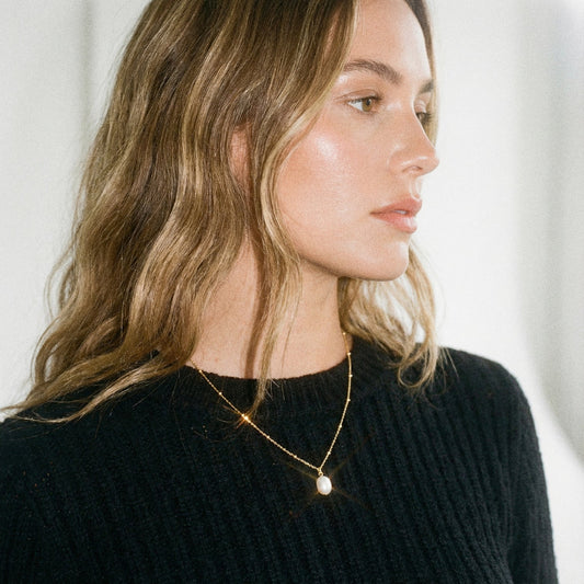 Woman wearing a black sweater and gold necklace with a pearl pendant against a white background